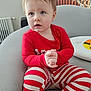 Meradith is registered to the contest to win money with this photo: toddler, child, red_clothing, striped_pants, sitting, armchair, indoor, person, cute, young_child, hands_clasped, light_gray_furniture, curious_expression, casual_wear, home_interior, plant, radiator, plush, portrait, baby