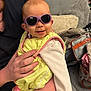 baby, infant, child, sunglasses, smile, hand, adult_hand, bib, onesie, couch, pillow, blanket, living_room, portrait, person, accessory, bag, zipper, fabric, cozy