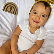 Ariel is registered to the contest to win money with this photo: baby, infant, smiling, white_onesie, headband, yellow_bow, milestone_block, 9_months, wooden_block, blanket, soft_texture, happy, portrait, cute, child, sitting, hands, bracelet, indoors, closeup