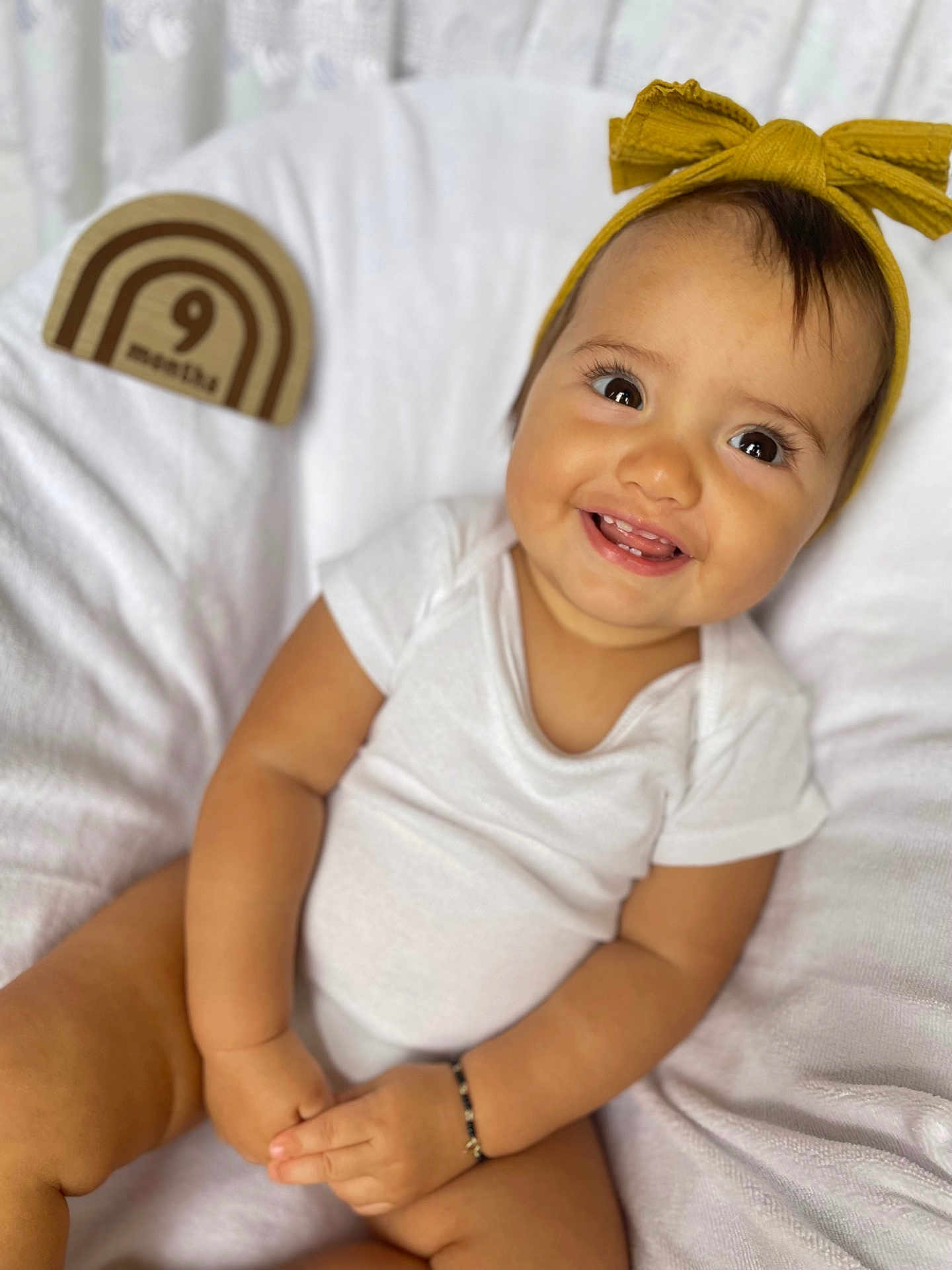 Ariel is registered to the contest to win money with this photo: baby, infant, smiling, white_onesie, headband, yellow_bow, milestone_block, 9_months, wooden_block, blanket, soft_texture, happy, portrait, cute, child, sitting, hands, bracelet, indoors, closeup