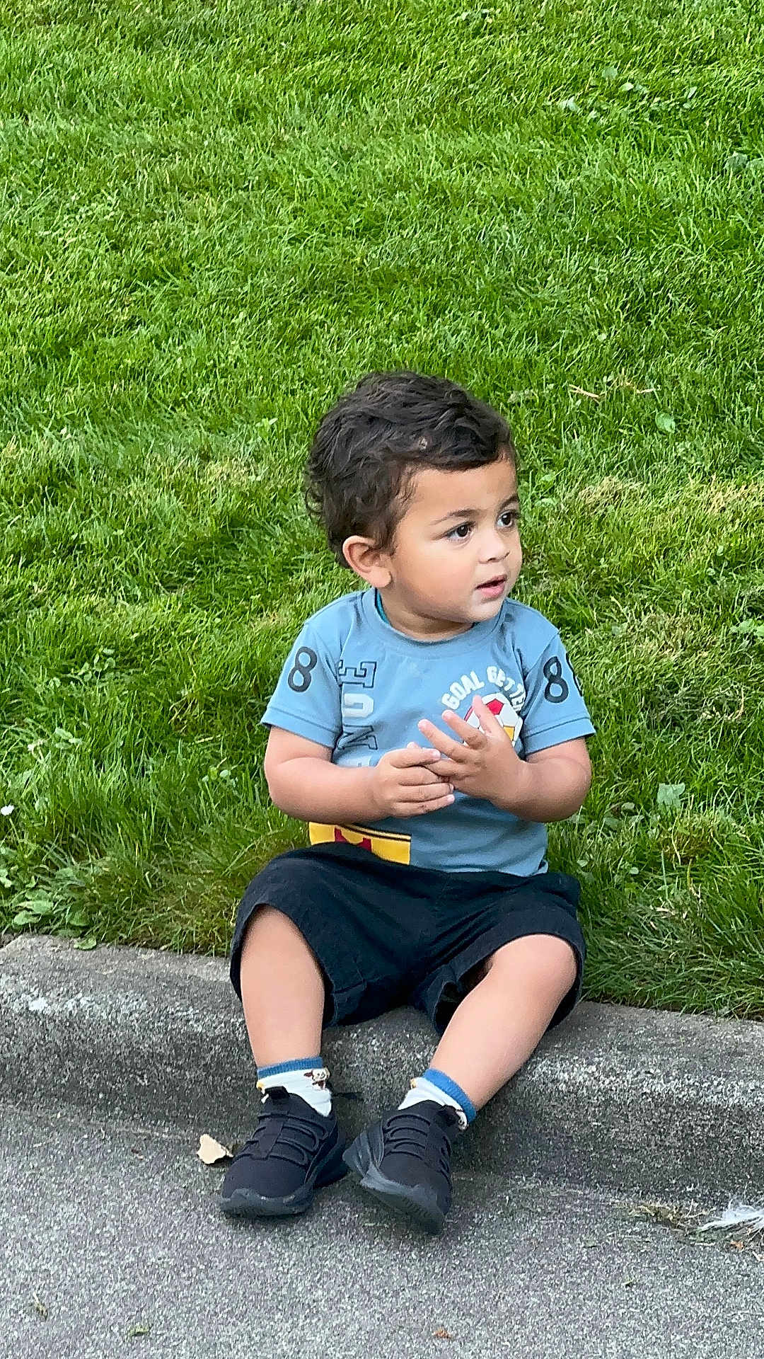 Avyaan joined the competition — help win amazing prizes! child, toddler, grass, sidewalk, curb, blue_shirt, black_shorts, sneakers, socks, outdoor, playing, sitting, young_child, casual_clothing, nature, greenery, daylight, cute, portrait, curious