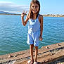child, girl, smiling, peace_sign, water, lake, outdoor, blue_sky, romper, sandals, rocks, sunlight, summer, nature, happy, standing, casual_clothing, daytime, barefoot, vacation