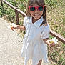 casual_wear, child, fence, flower, girl, greenery, hair, happy, nature, outdoor, person, playful, sandals, shadow, sidewalk, smile, summer, sunglasses, sunlight, white_dress