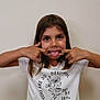 child, girl, funny_face, tongue_out, white_tshirt, floral_design, text_on_clothing, brown_hair, indoor, playful, portrait, smiling_eyes, casual_clothing, human, person, cute, childhood, expression, young, happy
