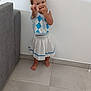 toddler, child, barefoot, pointing, smiling, indoor, floor, tile_floor, skirt, top, hair_tied, white_wall, couch, gray_couch, playful, person, young_child, skin, foot, hand
