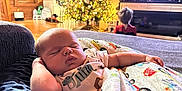 Wayne is registered to the contest to win money with this photo: baby, sleeping, blanket, couch, living_room, christmas_tree, holiday, child, television, furniture, wood_floor, decorations, indoors, cozy, relaxing, person, patterned_clothing, home, resting, soft_lighting