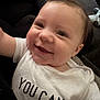 baby, smiling, close_up, infant, white_shirt, text_on_clothing, happy, face, child, young, cute, portrait, indoors, person, skin, eyes, mouth, hair, expression, toddler