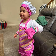 Aya joined the competition — help win amazing prizes! child, chef_hat, oven_mitt, apron, pink, striped_pants, barefoot, indoor, living_room, sofa, carpet, table, blanket, smiling, person, home, playful, cute, young_child, girl