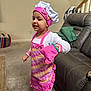child, chef_hat, oven_mitt, apron, pink, striped_pants, barefoot, indoor, living_room, sofa, carpet, table, blanket, smiling, person, home, playful, cute, young_child, girl