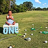 baby, child, birthday, outdoor, grass, golf_club, golf_ball, putting_green, crate, blue_sky, trees, sunny, smiling, first_birthday, celebration, playful, greenery, happy, sitting, toy