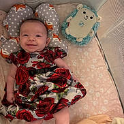 Natalia is registered to the contest to win money with this photo: baby, infant, smiling, dress, red_roses, pillow, fox_pattern, plush_toy, blue_plush, blanket, floral_pattern, playpen, mesh_sides, soft_bedding, indoor, cute, happy, child, laying_down, cozy