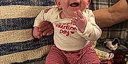 Natalia is registered to the contest to win money with this photo: baby, infant, smiling, happy, sitting, clothing, striped_pants, long_sleeve_shirt, valentine_day, pillow, blanket, couch, hand, adult, indoors, cozy, cheerful, face, person, home