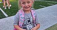 Presley is registered to the contest to win money with this photo: child, girl, smiling, outdoor, football_field, sports_field, backpack, bow, blonde_hair, portrait, standing, school, stadium, spectators, grass, artificial_turf, daylight, sports, casual_clothing, happy