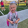 child, girl, smiling, outdoor, football_field, sports_field, backpack, bow, blonde_hair, portrait, standing, school, stadium, spectators, grass, artificial_turf, daylight, sports, casual_clothing, happy