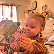 Hélèna a rejoint le concours — aidez-le/la à gagner de superbes lots ! baby, blanket, board_book, book, child, clock, cozy, cute, dresser, face, hair_tie, hand, indoor, living_room, pigtails, portrait, reading, smile, toddler, toy