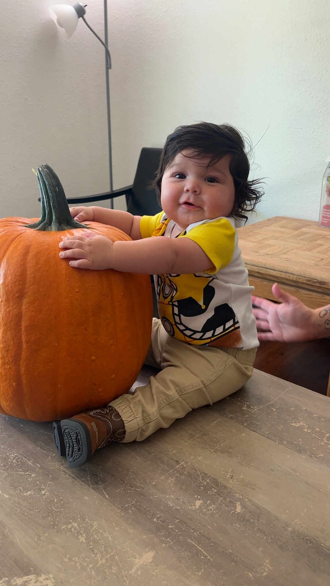 Abel is registered to the contest to win money with this photo: baby, child, pumpkin, table, indoor, yellow_shirt, beige_pants, boots, dark_hair, cute, smiling, hand, chair, lamp, wooden_table, person, sitting, fall, autumn, home
