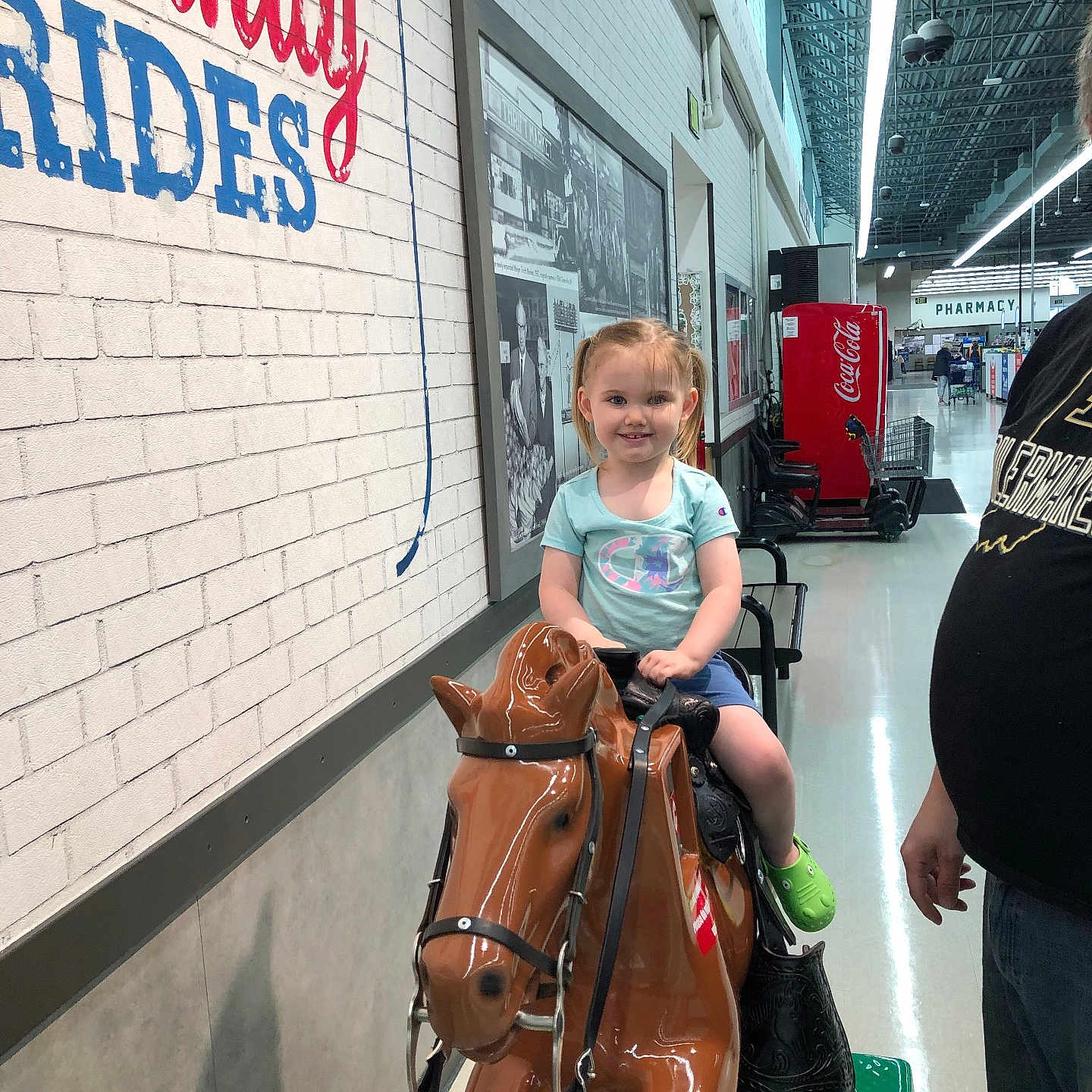 Nedra is registered to the contest to win money with this photo: aisle, brick_wall, casual_clothing, child, coin_operated_ride, fun, girl, green_shoes, happy, horse, indoor, pigtails, playful, portrait, shopping_area, signage, smiling, store, toy, vending_machine