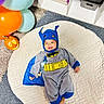 baby, balloons, batman, blue, cape, carpet, carved, child, costume, cute, face, floor, gray, happy, indoors, infant, mat, pumpkin, smile, toy