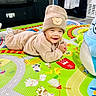 animal_prints, baby, barn, beige_clothing, carpet, child, colorful, cozy, cute, floor, happy, hat, indoor, infant, play_mat, plush, portrait, smiling, toddler, toy