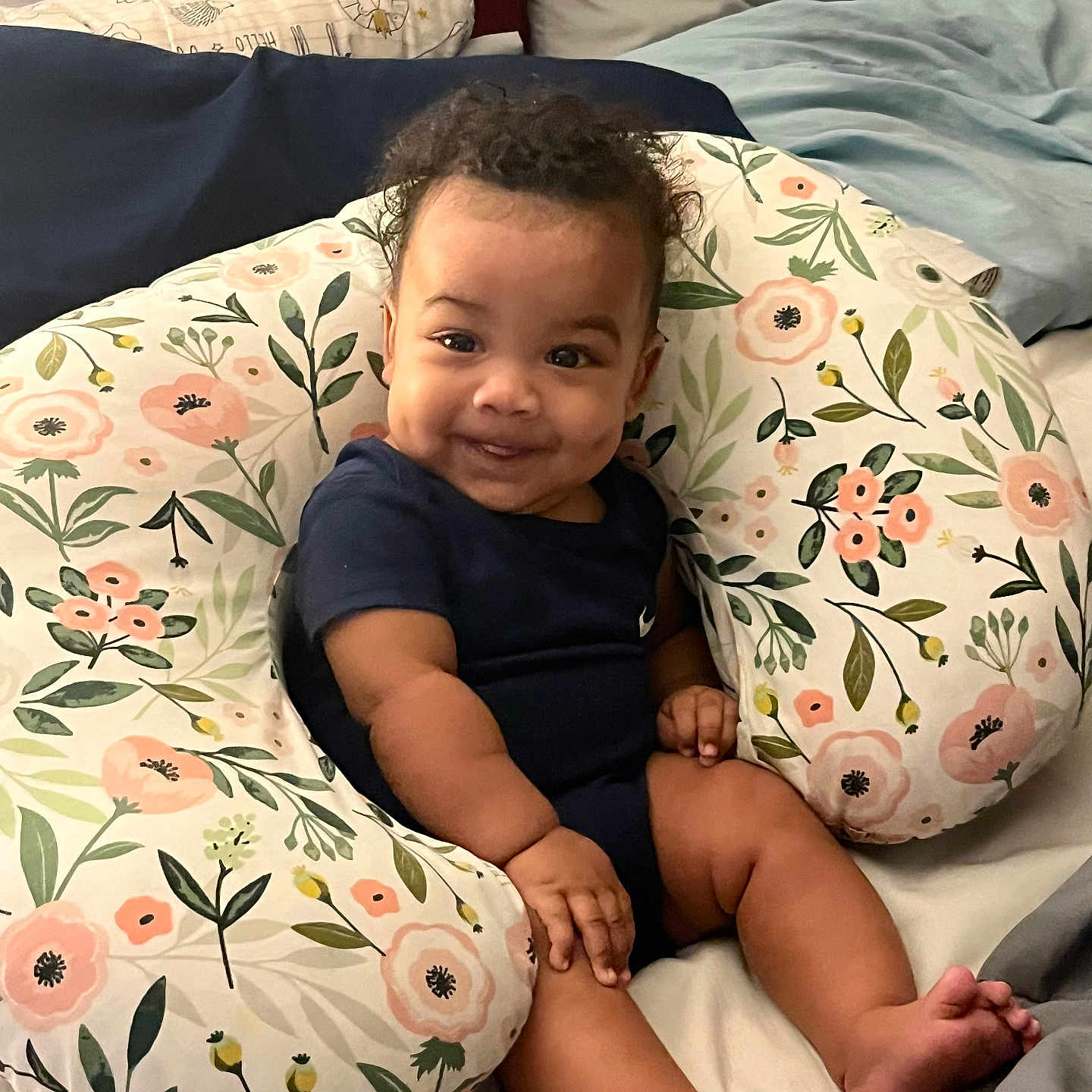 Janarious is registered to the contest to win money with this photo: baby, infant, smile, curly_hair, floral_pillow, bedding, bed, comfortable, cute, child, person, indoors, happy, skin, face, legs, hand, toe, blue_clothing, cozy