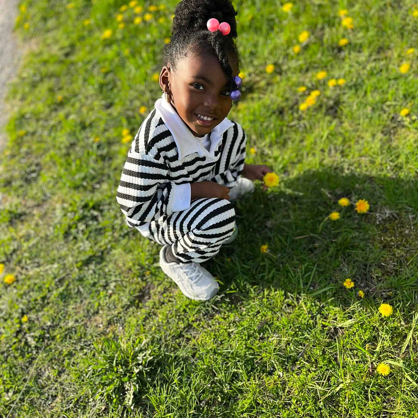 Kai joined the competition — help win amazing prizes! child, girl, smiling, outdoor, grass, flowers, yellow_flowers, sunlight, crouching, hair_beads, striped_clothing, casual_wear, white_shoes, nature, greenery, happy, portrait, daylight, young, person