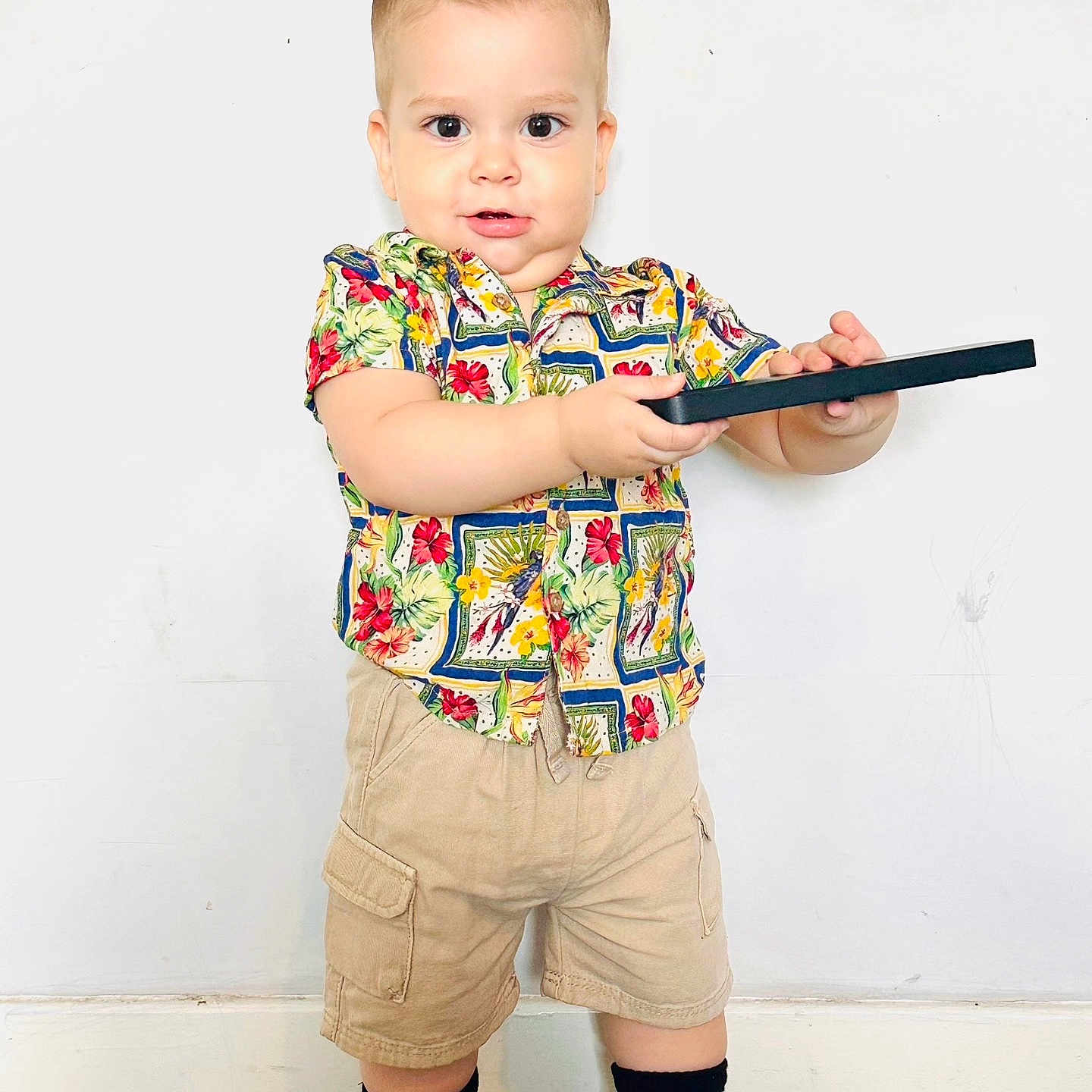 Matias joined the competition — help win amazing prizes! beige_shorts, black_object, boy, carpet, casual_clothing, child, colorful_shirt, curious, cute, feet, floral_pattern, hands, indoor, playful, portrait, socks, standing, toddler, white_wall, young_child