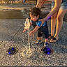 Isaiah joined the competition — help win amazing prizes! toddler, child, water_fountain, water, adult, hand, legs, outdoor, sunlight, pavement, curiosity, play, people, clothing, shoes, support, daylight, public_space, fun, interaction