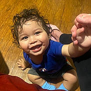 Isaiah joined the competition — help win amazing prizes! toddler, child, smiling, curly_hair, blue_shirt, socks, wooden_floor, happy, indoor, person, cute, baby, hand, sitting, playful, young_child, portrait, casual_clothing, bright, curious