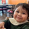 Isaiah is registered to the contest to win money with this photo: toddler, child, smile, messy_hair, bib, food_stain, table, restaurant, chair, orange_wall, indoor, happy, person, young_child, casual, seated, cute, face, hands, curly_hair