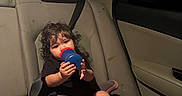 Isaiah is registered to the contest to win money with this photo: toddler, child, car_seat, sippy_cup, curly_hair, car_interior, backseat, seatbelt, toy, footwear, sock, dim_lighting, seat, vehicle, candid, sitting, young_child, indoors, casual, relaxed