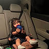 toddler, child, car_seat, sippy_cup, curly_hair, car_interior, backseat, seatbelt, toy, footwear, sock, dim_lighting, seat, vehicle, candid, sitting, young_child, indoors, casual, relaxed
