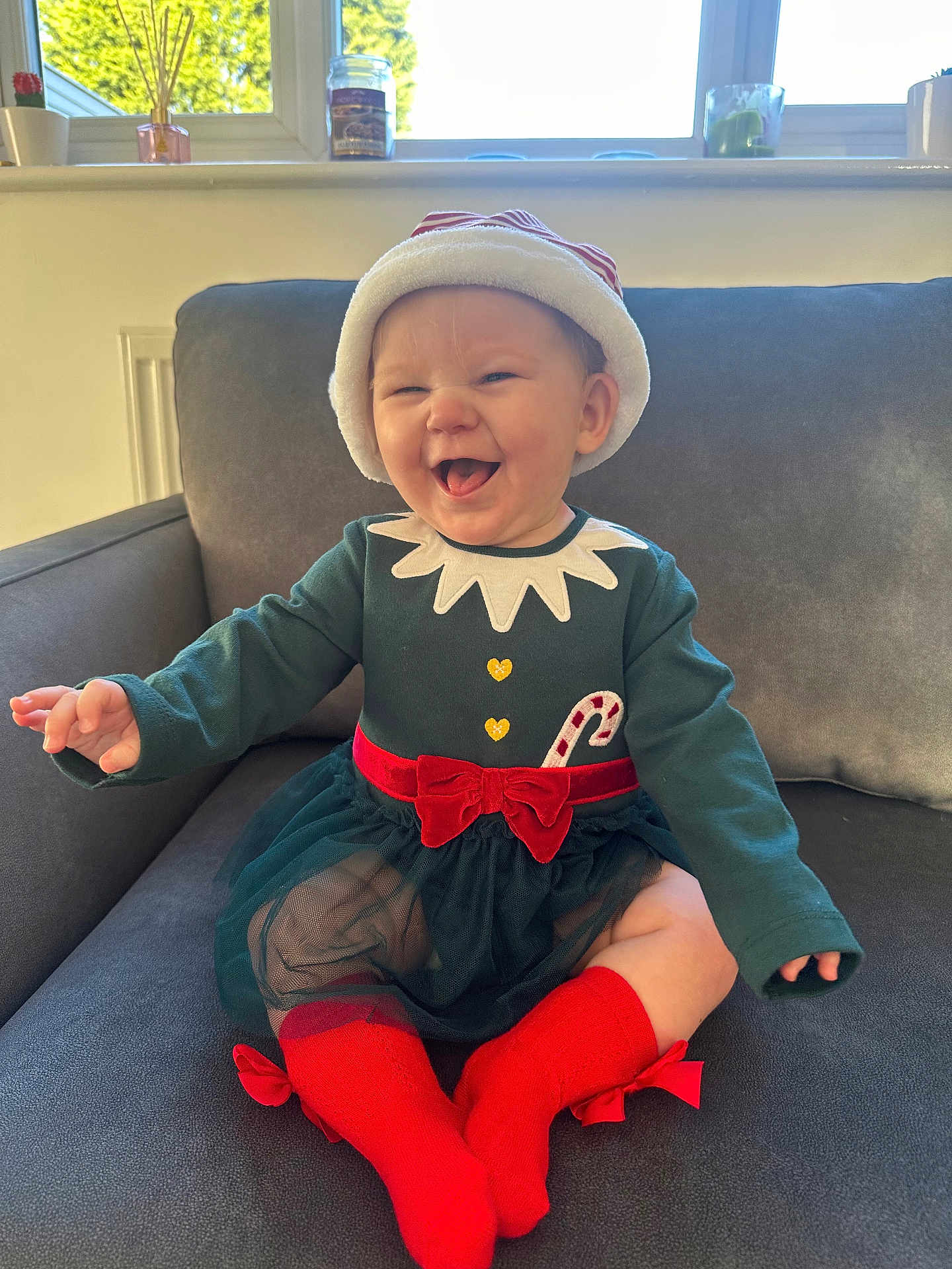 Nova-Mae is registered to the contest to win money with this photo: baby, child, smiling, holiday_outfit, santa_hat, candy_cane, red_socks, green_dress, bow, couch, indoor, window, plants, happy, cute, portrait, toddler, festive, cozy, celebration