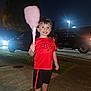 child, boy, cotton_candy, red_shirt, black_shorts, sidewalk, night, streetlights, cars, grass, smile, short_hair, outdoor, holding, food, happy, casual_clothing, person, recreation, evening