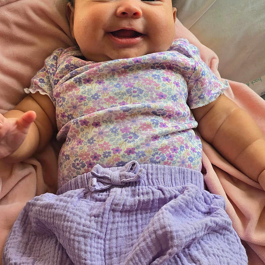 Arisbeth is registered to the contest to win money with this photo: baby, bed, blanket, candid, child, cute, face, floral_clothing, hands, happy, home, indoor, infant, lavender_shorts, legs, lying_down, photography, pillow, portrait, smiling