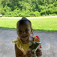 Mahanna is registered to the contest to win money with this photo: child, toddler, yellow_dress, smiling, holding, beverage_pouch, outdoor, grass, trees, concrete, sunny, happy, person, young_child, nature, summer, casual_clothing, daylight, front_view, cute