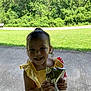 child, toddler, yellow_dress, smiling, holding, beverage_pouch, outdoor, grass, trees, concrete, sunny, happy, person, young_child, nature, summer, casual_clothing, daylight, front_view, cute