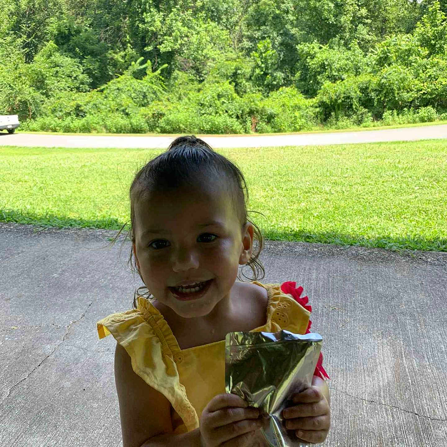 Mahanna is registered to the contest to win money with this photo: beverage_pouch, casual_clothing, child, concrete, cute, daylight, front_view, grass, happy, holding, nature, outdoor, person, smiling, summer, sunny, toddler, trees, yellow_dress, young_child