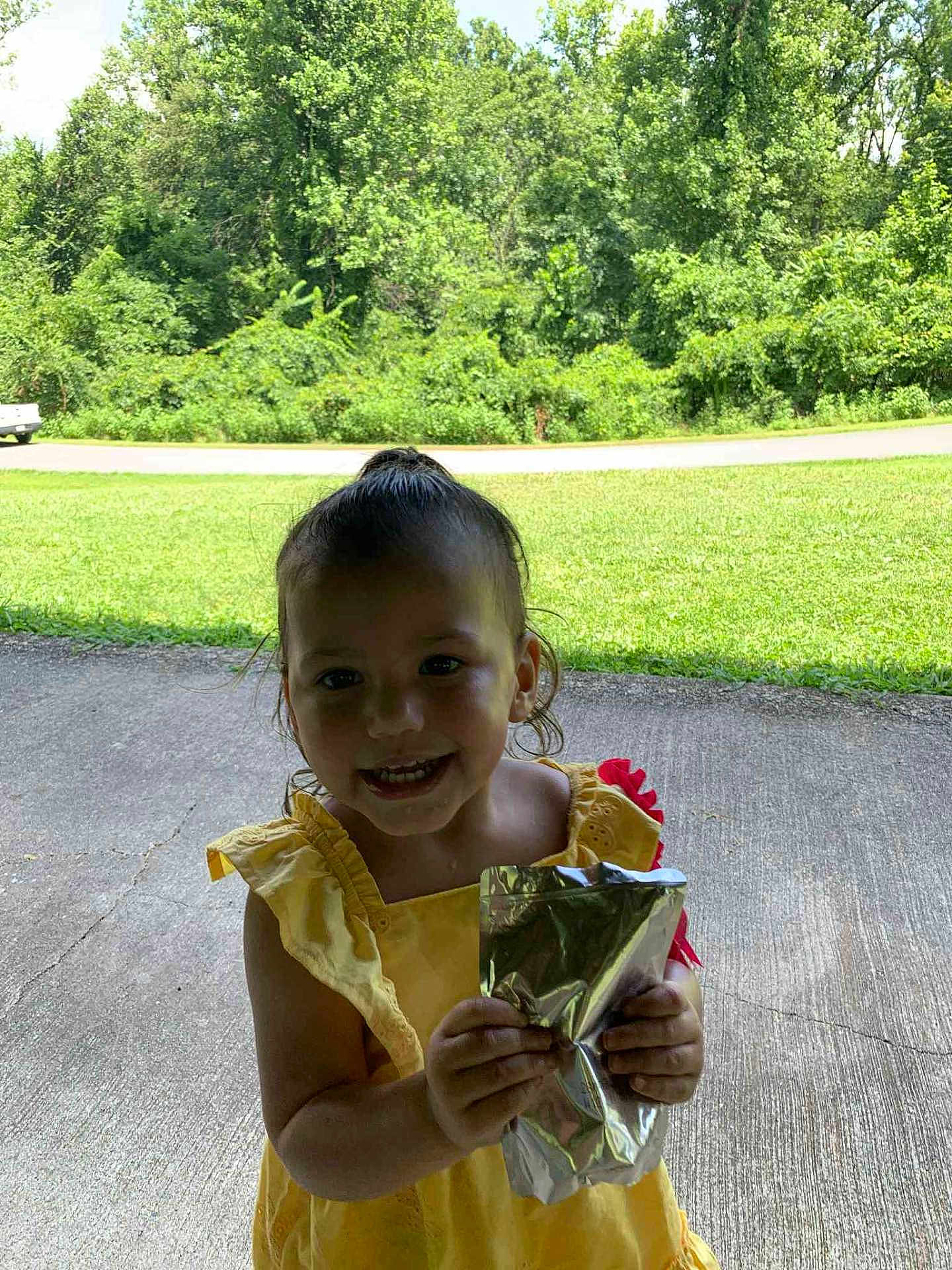 Mahanna is registered to the contest to win money with this photo: child, toddler, yellow_dress, smiling, holding, beverage_pouch, outdoor, grass, trees, concrete, sunny, happy, person, young_child, nature, summer, casual_clothing, daylight, front_view, cute