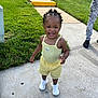 toddler, child, smiling, outdoor, sidewalk, grass, yellow_clothing, white_shoes, steps, hairstyle, person, concrete, sunlight, casual_wear, happy, young_child, greenery, summer, urban, playful