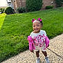 child, toddler, smiling, girl, pink_bows, pink_jacket, white_shirt, white_shorts, sneakers, sidewalk, grass, brick_building, outdoor, happy, cute, portrait, daylight, person, young_child, fashion