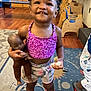 toddler, child, smiling, indoor, doll, toy, purple_top, floral_shorts, carpet, wooden_floor, playroom, furniture, person, footwear, pink_shoes, happy, cute, portrait, girl, braided_hair