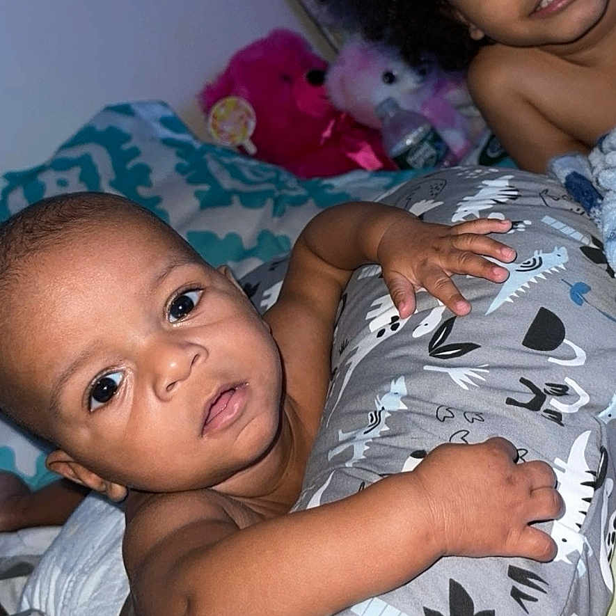 Kyius is registered to the contest to win money with this photo: baby, bedroom, blanket, child, close_up, cozy, curly_hair, cute, dinosaur_pattern, face, indoor, lying_down, person, pillow, playful, skin, smiling, stuffed_animals, toddler, toy