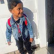 Aiden joined the competition — help win amazing prizes! child, boy, denim_jacket, backpack, sneakers, outdoor, sidewalk, shadow, white_fence, smiling, happy, casual_clothing, sunlight, daytime, portrait, standing, black_pants, red_straps, short_hair, playful
