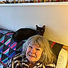woman, cat, black_cat, quilt, couch, shelf, bird_figurines, indoor, smiling, robe, gray_hair, seated, home_decor, colorful, furniture, pet, happy, portrait, relaxed, casual