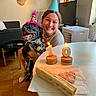 woman, dog, party_hat, birthday_candles, table, treats, celebration, smiling, indoor, floor, sofa, lamp, poster, rainbow_flag, window, radiator, television, happy, pet, friendship