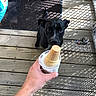 dog, black_dog, ice_cream_cone, hand, wooden_deck, shoes, shadow_pattern, keys, person, outdoor, summer, waiting, curious, pet, leisure, sunlight, footwear, ground, holding, animal