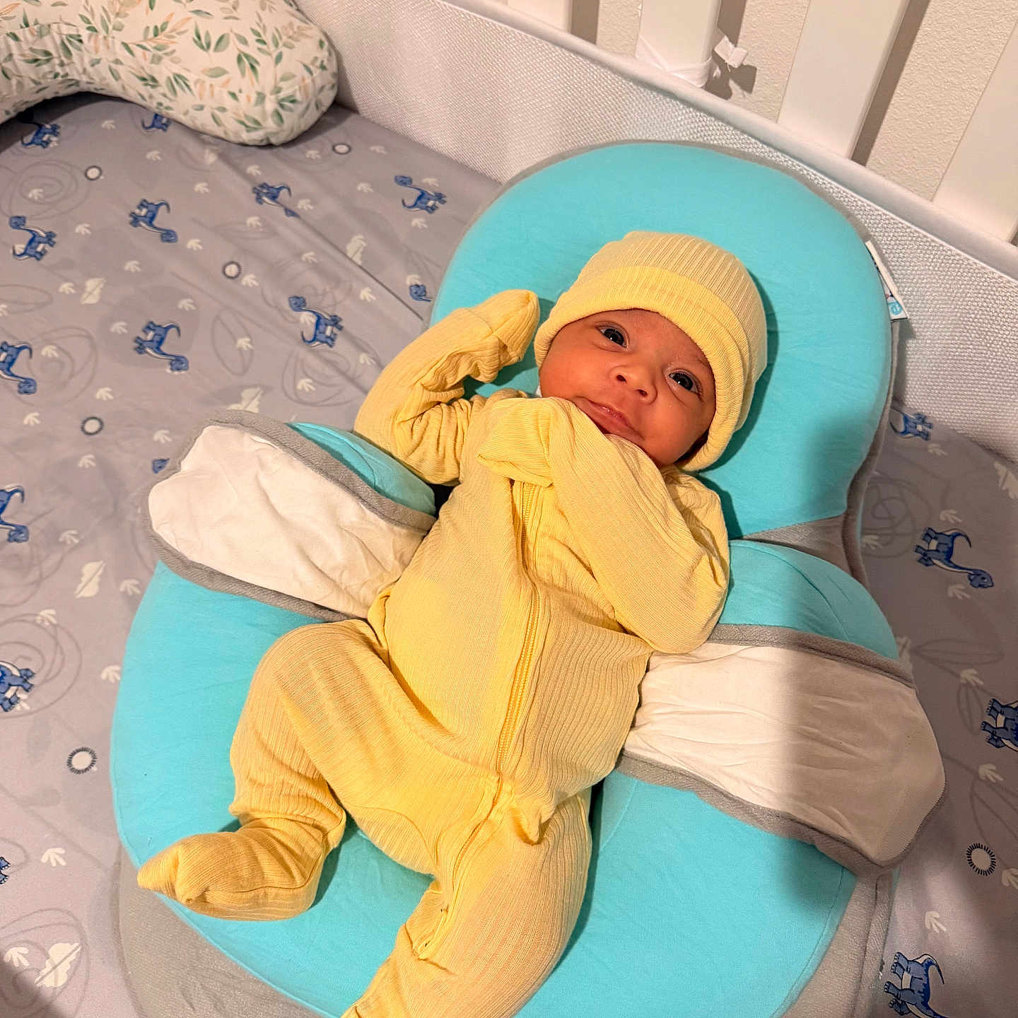 Mateo is registered to the contest to win money with this photo: baby, newborn, child, infant, yellow_outfit, knit_cap, smiling, awake, lying_down, baby_nest, turquoise, crib, nursery, mattress, pillow, blanket, patterned_fabric, portrait, indoor, cozy