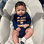 baby, child, infant, onesie, tongue_out, baby_seat, cushion, toy, curly_hair, socks, playful, indoor, cute, smiling, person, young_child, comfortable, colorful_toy, text_on_clothing, relaxed