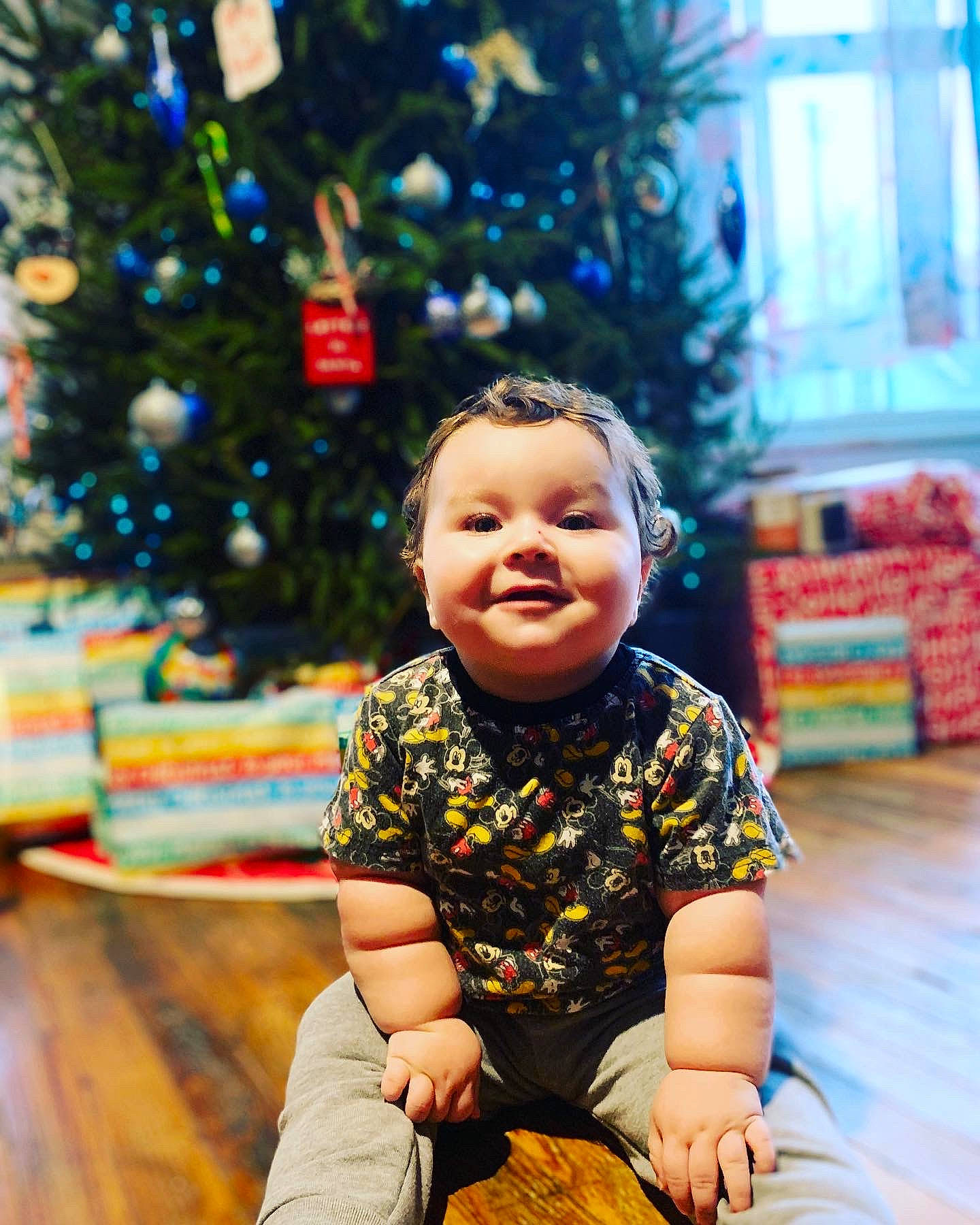 Eli is registered to the contest to win money with this photo: child, christmas_decoration, christmas_ornament, christmas_tree, event, eye, flooring, fun, hairstyle, happy, human_body, leisure, people_in_nature, person, plant, sitting, skin, smile, standing, toddler
