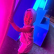 Takari is registered to the contest to win money with this photo: child, toddler, hood, smile, barefoot, neon_lights, pink_light, blue_light, night, glowing, curious, standing, outdoor, playful, colorful, person, clothing, texture, joyful, illumination